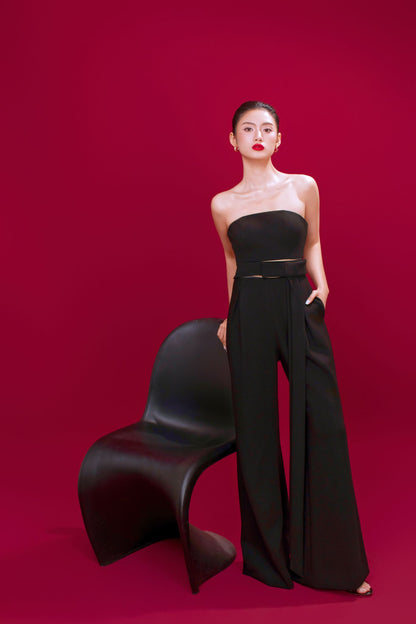 Woman in a black strapless top and high-waisted pants standing next to a modern black chair on a red background